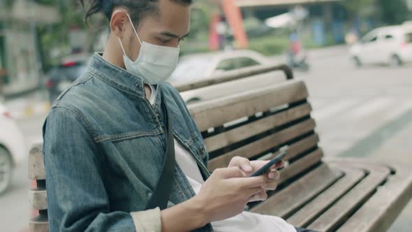 Asian man wearing a mask and using a smartphone while sitting on a bench.