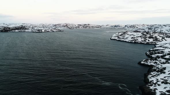 The Drone Rises Above the Dark Waters of the Arctic Ocean Near the Rocky Shore Covered with Snow