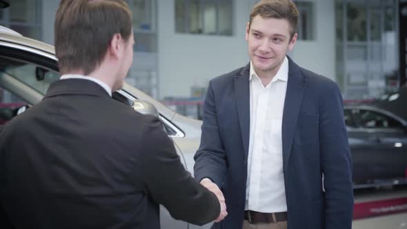 Portrait of Thankful Caucasian Man Shaking Dealer's Hand in Car Dealership alt