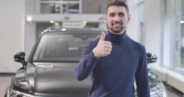 Portrait of Young Caucasian Man Standing in Front of New Car and Showing Thumb Up alt