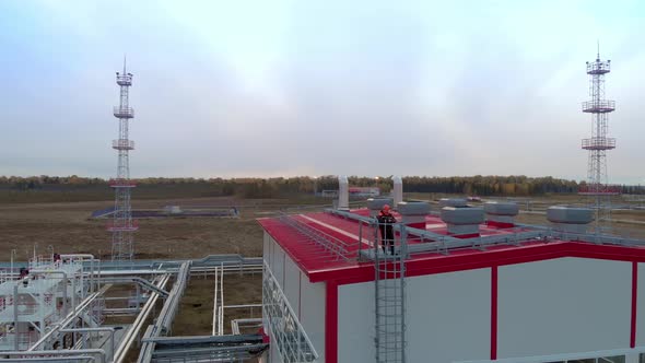 A Drone Flies an Oil and Gas Worker in an Orange Helmet and Overalls Who Stands on the Roof of a Gas alt