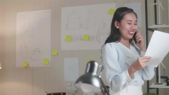 Asian Woman Designer Talking On Smartphone While Looking At The Layout Bond In Hand And Walking alt