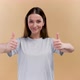 Young Cheerful Woman Shows Ok Sign Thumbs Up Looking at the Camera Ob a Beige Background - VideoHive Item for Sale