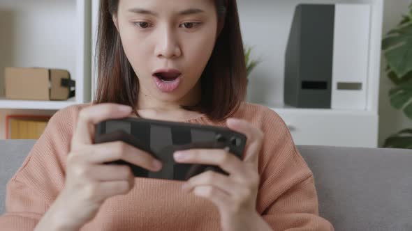 Excited young Asian woman playing games on smartphone in the living room at home. alt