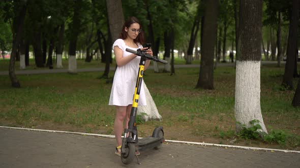 Young Asian Woman in a White Dress Starts a Ride on a Sharing Electro Scooter in the Park alt