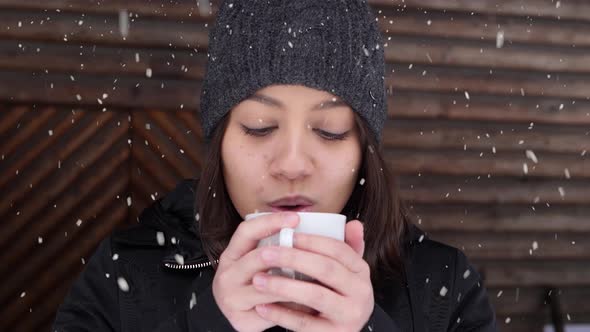 Attractive Asian female drinking hot beverage while outdoors against wooden wall during snowfall alt