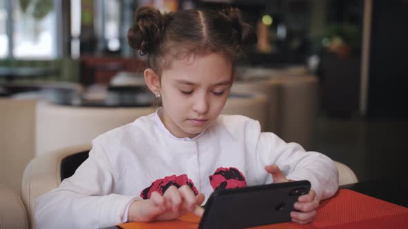 Little Girl Can Not Break Away From the Smartphone Sitting at a Table in a Cafe alt