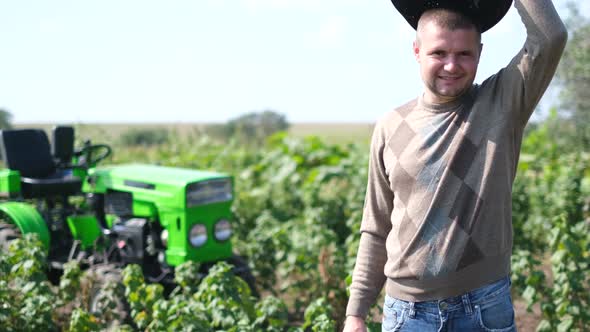 Young Farmer in a Hat Standing on the Background of a Tractor and Smiling at the Camera alt