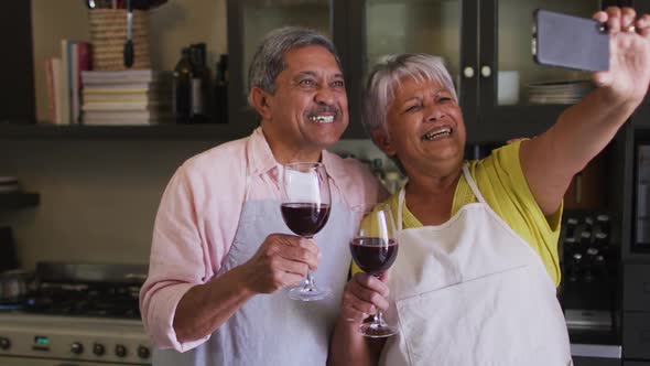 Happy senior mixed race couple taking selfie toasting with wine in kitchen alt