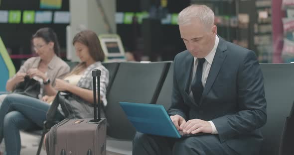 Serious Mid-adult Caucasian Businessman Using Laptop in Airport Waiting Area with People Talking at alt
