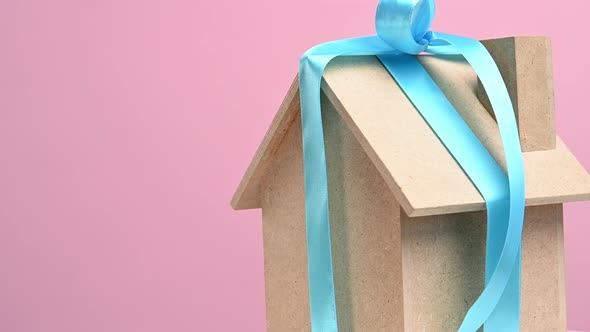model of a wooden house tied with a blue silk ribbon on a pink background alt