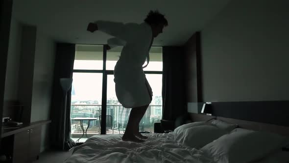 Happy Young Man Jumping on Bed at Hotel in Bathrobe alt
