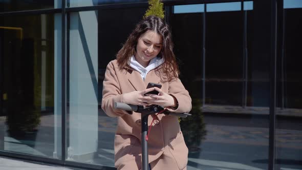 A Young Student Girl in a Sweater Jeans and a Coat Stands with an Electric Scooter Near a Stylish alt