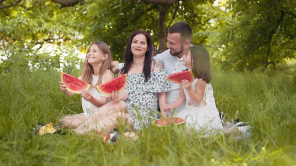 Cheerful Parents with Daughters in Summer Outfits Sitting Together at Green alt