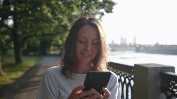 a Woman Carefully Looks at the Phone Screen and Smiles on the City Embankment alt