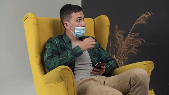 Man Sits in a Chair and Uses a Phone with a Medical Mask  and After Being Asked