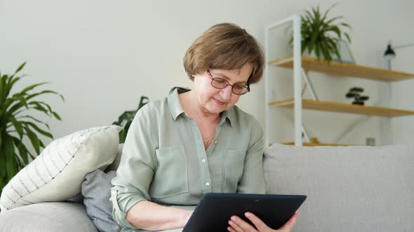 Smiling Elderly Woman Resting on Sofa Using Digital Tablet at Home alt