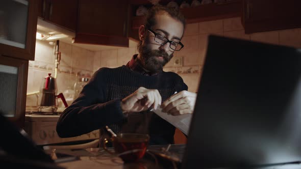 Young Hipster Bearded Man with Glasses Is Opens a Invoice and Pays Invoice Online in Kitchen