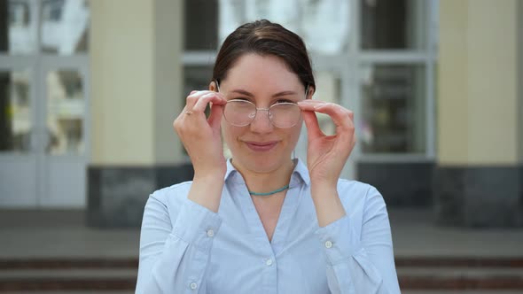 A Young Girl Stands Against the Background of the University and Puts on Glasses alt