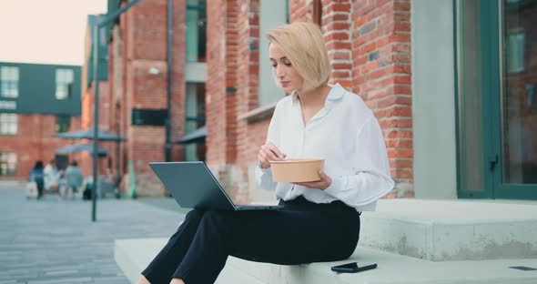 Woman Having Vegetables Salad for Lunch, Sitting on Stairs During Using Laptop alt