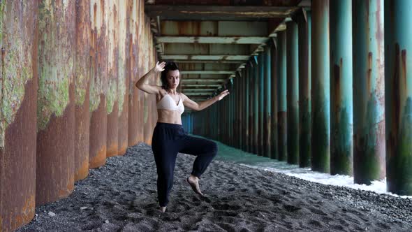 a Barefoot Woman Dances Under the Pillars of the Bridge Against the Background of the Incoming Waves alt