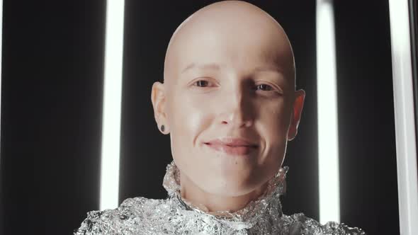 Young Bald Woman in Foil Wear Laughing at Camera in Dark Studio alt