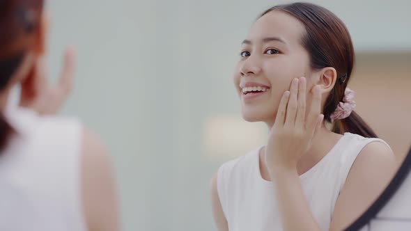 Fresh healthy skin of Asian girl looking at mirror, applying facial moisturizer on her face. alt