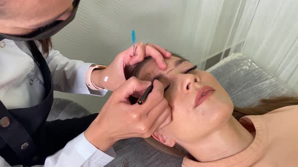 Beauty Master in a Mask Draws a Contour of Eyebrows on a Young Girl