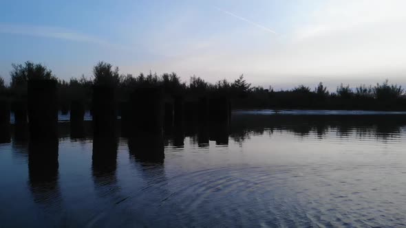 Silhouetted Trees Sit on the Bank of a Tranquil River, Revealing an Industrial City and Arch Bridge alt