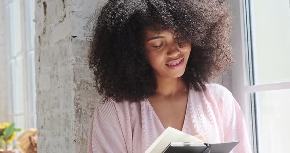 A Thoughtful Black Young Woman with Curly Hair Looks Around and Writes in a Paper Notebook Near the alt
