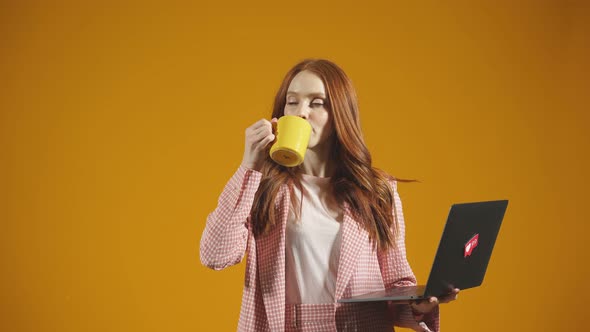 Busy Young Woman with a Laptop and a Mug Stands Against an Isolated Orange Background in the Studio