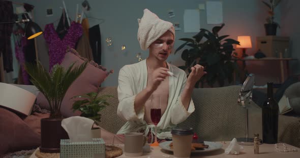 Front View of Man Transgender in Bath Robe Sitting on Sofa and Drying Fingernail Polish. Man Trans alt