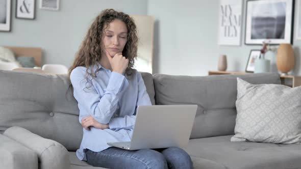 Curly Hair Woman Thinking and Working on Laptop While Sitting on Couch alt