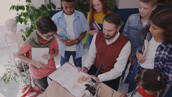 Male Teacher Explaining Topic in Biology Textbook to Children Surrounding Him alt