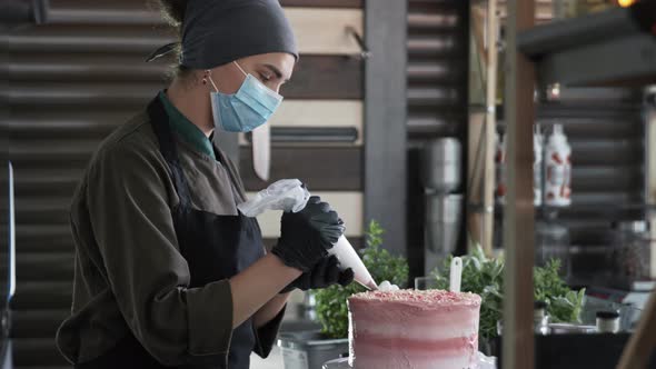 Female Baker Uses Precautions in a Medical Mask and Gloves Decorated Homemade Sweet Cake with Cream alt