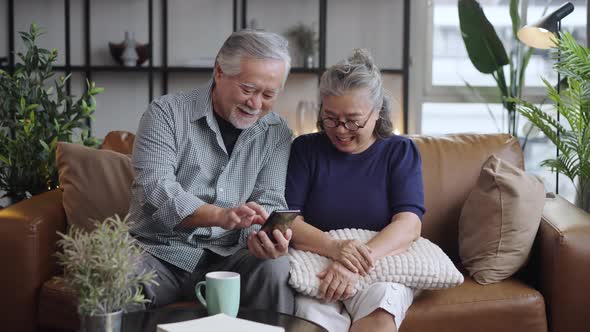 Asian mature senior couple using smart phone together and smiling on sofa at home