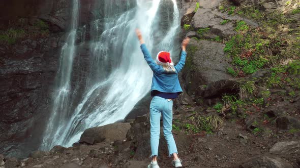 A girl in a Santa Claus hat and dances against the backdrop of a waterfall alt