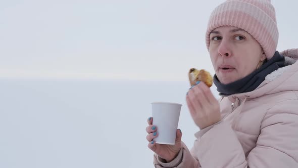 Portrait Young Woman on the Shore of the Frozen Sea Eats a Bun and Drinks Coffee alt