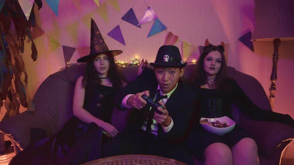 A Guy in a Creepy Costume is Actively Playing a Video Game Console and the Girls are Sitting and alt