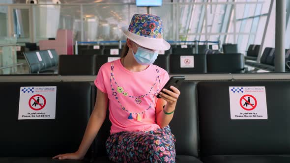 Teenage Girl, in Protective Mask, Uses Mobile. She Sitting in an Empty Airport Lounge, Waiting To alt