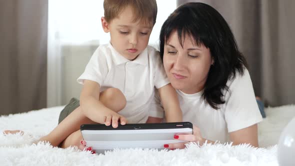 The Mother and the Little Boy Are Resting Lying on the Bed. The Child Plays with a Tablet alt
