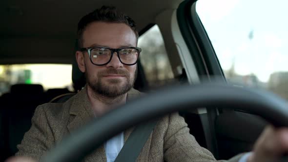Satisfied Bearded Man in Glasses Driving a Car Down the Street in Sunny Weather alt