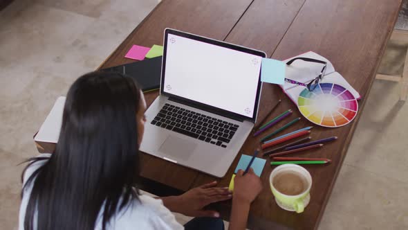 Mixed race woman using laptop writing in notebook drinking coffee working from home alt