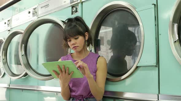 A Young Woman sits using her Digital Tablet whilst waiting for her laundry