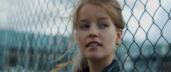 Young woman leaning towards metal fence and holding a straw in her mouth alt