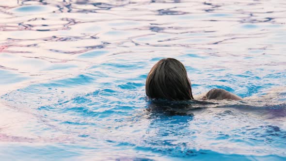 Woman Swims in a Pool with Clear Water on the Background of a Summer Sunset on Vacation alt