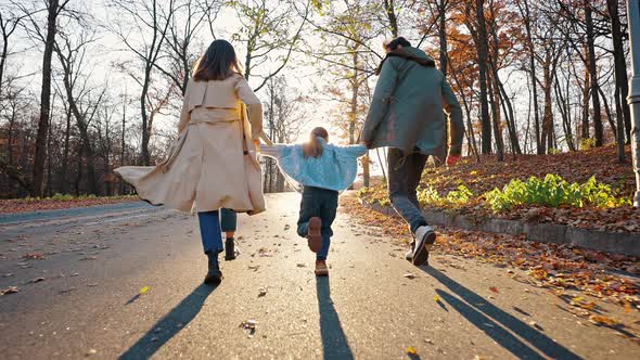 Unknown Young Parents Holding Hands of Little Daughter Playing Running By Paved Road in Autumn Park alt