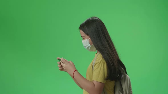 Girl Student Wearing A Mask And Playing Video Game On Mobile Phone While Walking On Green Screen alt