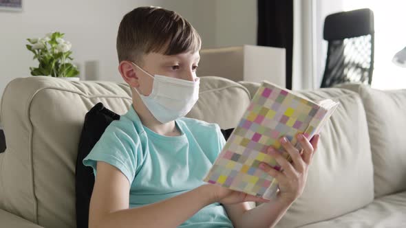 A Young Boy in a Face Mask Reads a Book As He Sits on a Couch at Home  alt