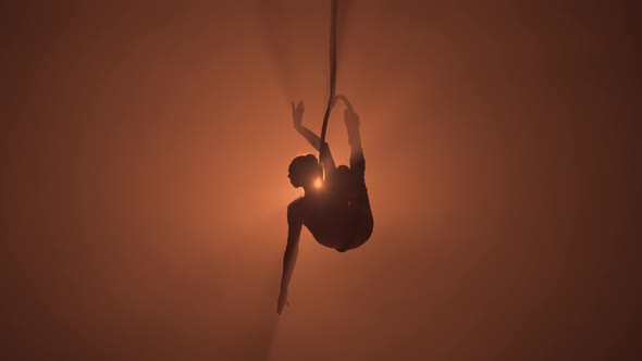 Silhouette of Flexible Aerial Gymnast Rotates on an Aerial Ring and Performs Acrobatic Tricks in the alt
